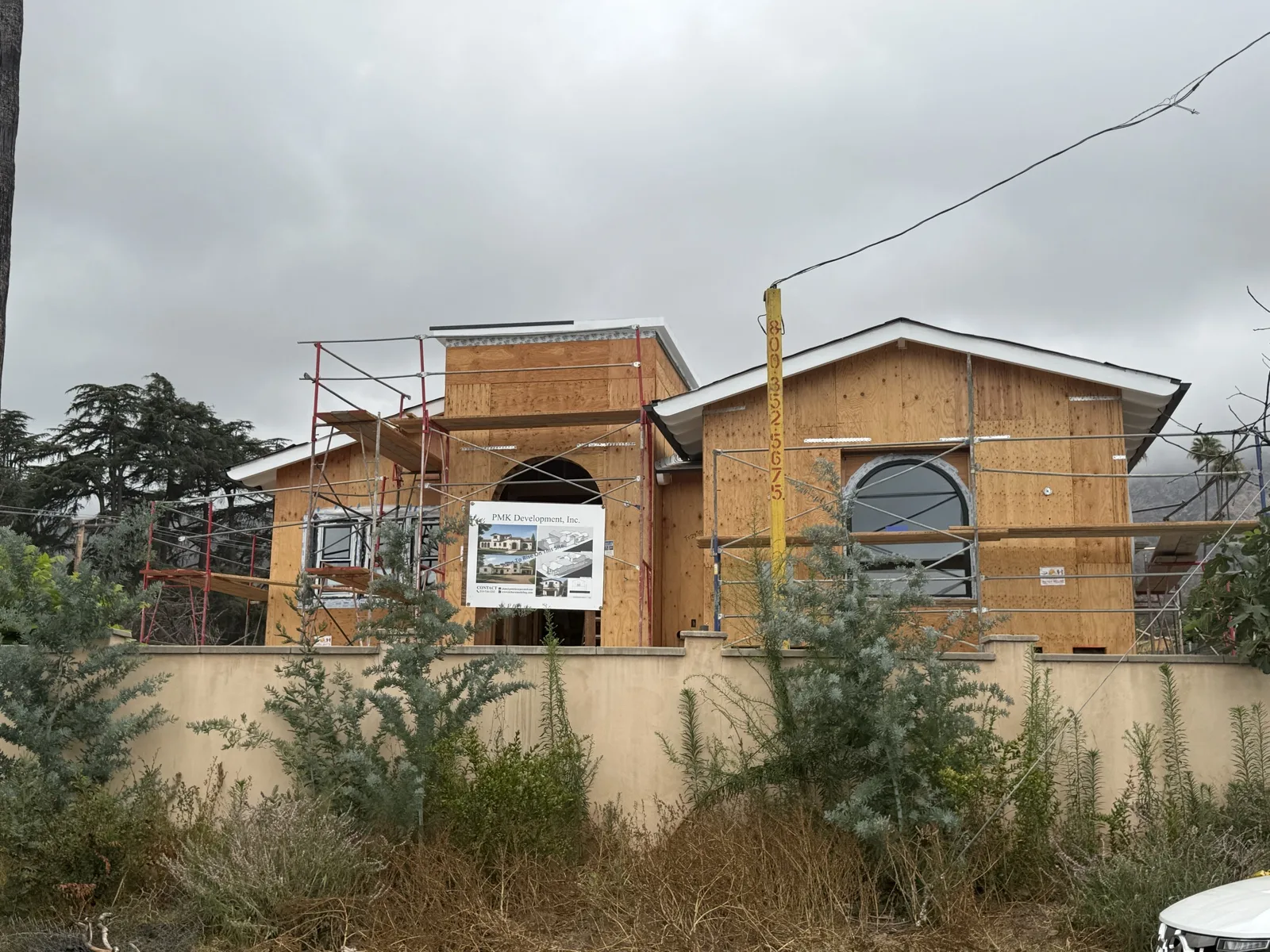 Front elevation of the Las Flores Drive rebuild behind its stucco perimeter wall: framed arched entry portico, arched window, and the PMK Development sign on the scaffold.